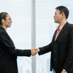 The head of a nonprofit wearing a long black coat shakes hands with a top donor in her office